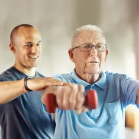 Senior resident working arms out with hand weight with rehab employee assisting.
