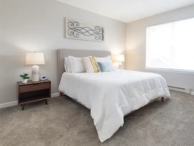 Senior living bedroom with bed, large window and side table with lamp, plant and alarm clock.