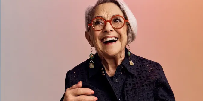 Older adult woman with short gray hair, wearing red glasses, dangling earrings, and a dark lacy top, smiles joyfully against a soft gradient background.
