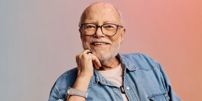 Smiling older man with glasses and beard, wearing a denim shirt and watch, against a soft gradient background.
