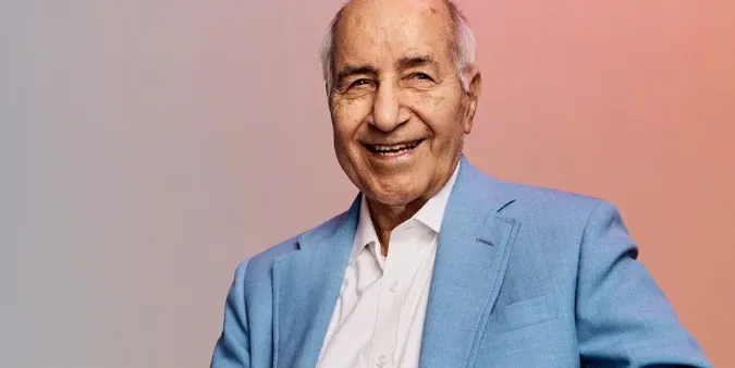 Older man smiling warmly, wearing a light blue suit and white shirt, with a soft gradient background.