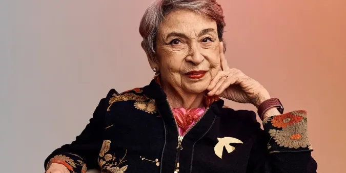 Older adult woman with short gray hair, wearing a floral-patterned jacket, holding red glasses, seated against a warm gradient background.