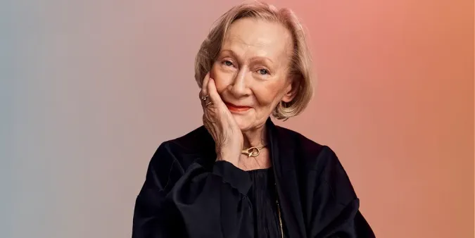 Older adult woman with short blonde hair smiles gently, resting her chin on her hand against a soft gradient background.