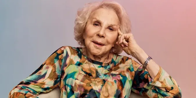 Older woman with blond hair, wearing a colorful blouse, sitting and smiling with her hand on her face against a gradient background.