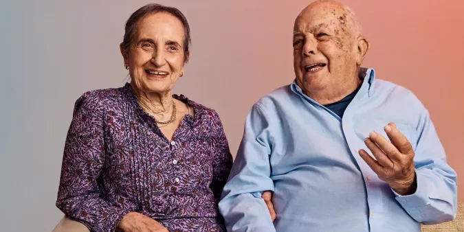 An older couple sitting together, the woman in a purple floral blouse smiles, while the man in a light blue shirt gestures animatedly.