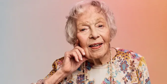 Older woman with short white hair, wearing a floral blouse, smiling gently, with her hand resting on her cheek against a soft gradient background.