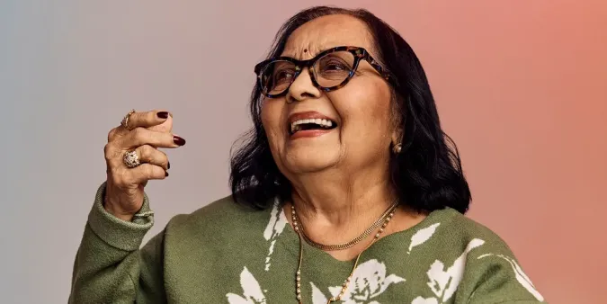 Smiling older woman with glasses, wearing a green floral sweater, holding a hand up against a gradient background.