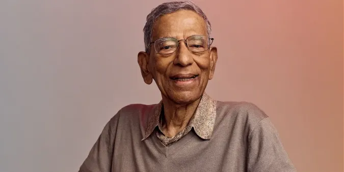 Older adult man with glasses and a warm smile, wearing a light gray sweater over a collared shirt, against a softly colored gradient background.