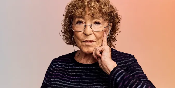 An older woman with curly hair and glasses, wearing a striped top, gestures thoughtfully with her hand on her chin against a soft gradient background.