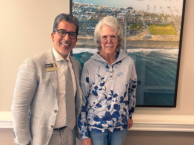Two people smiling, standing in front of a large aerial photograph of a coastal city. One wears a suit, the other a floral hoodie.