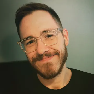A smiling man with a beard and clear glasses against a plain blue background.