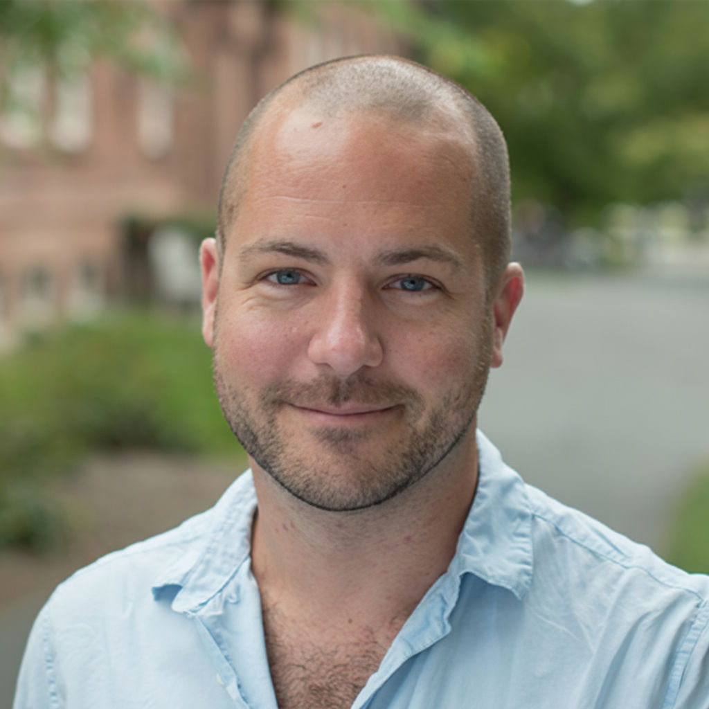 A man with a shaved head and light beard in a light blue shirt, smiling outdoors with a blurred background of trees and a building.