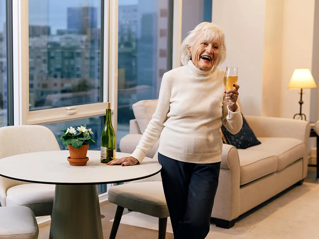 An older adult woman in a cozy apartment smiles while holding a glass of wine near a table with a potted plant and a bottle. City buildings visible outside.