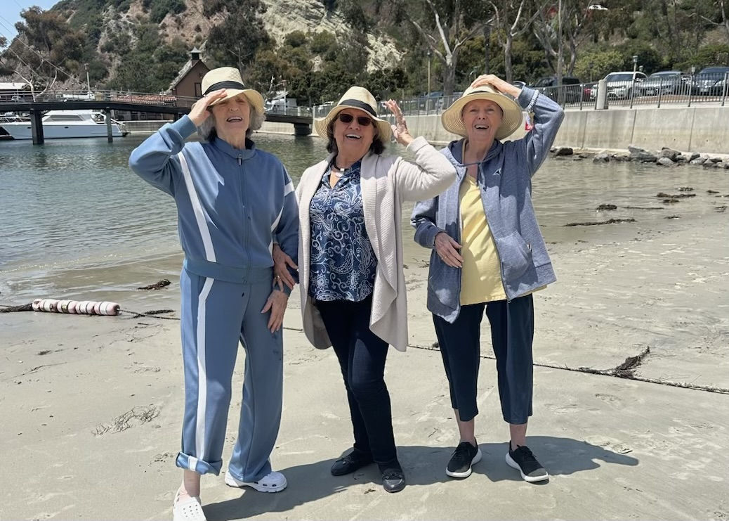 Three women in casual outfits and hats stand on a sandy beach, smiling and shielding their eyes from the sun. A pier and trees are in the background.