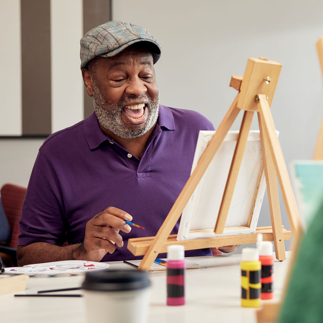 Older man in a purple shirt and cap joyfully painting at an easel, surrounded by colorful paint bottles and a coffee cup.