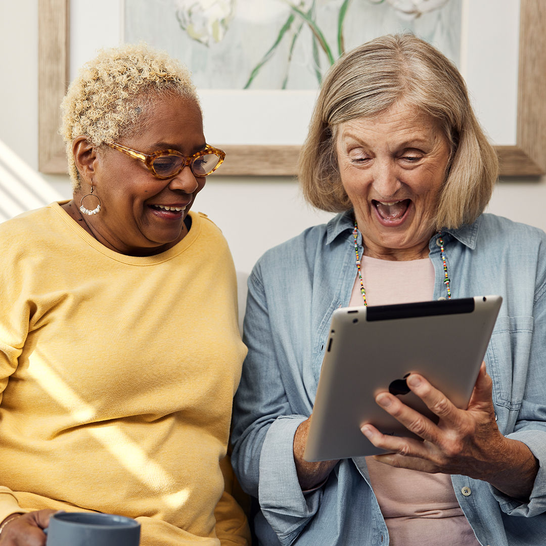 Two older women smiling, seated on a couch, looking at a tablet. One holds a mug. A framed floral picture hangs on the wall behind them.