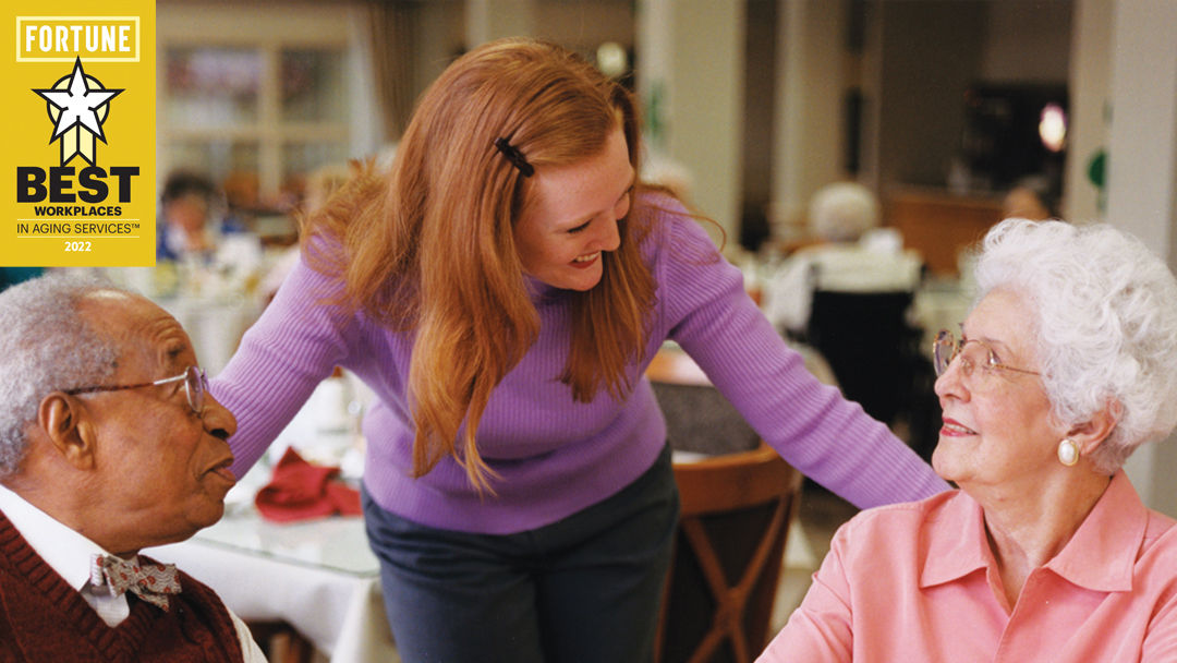 Staff member talking to two senior living residents while they eat in the dining room. Fortune Best Workplaces logo in the upper left hand corner.