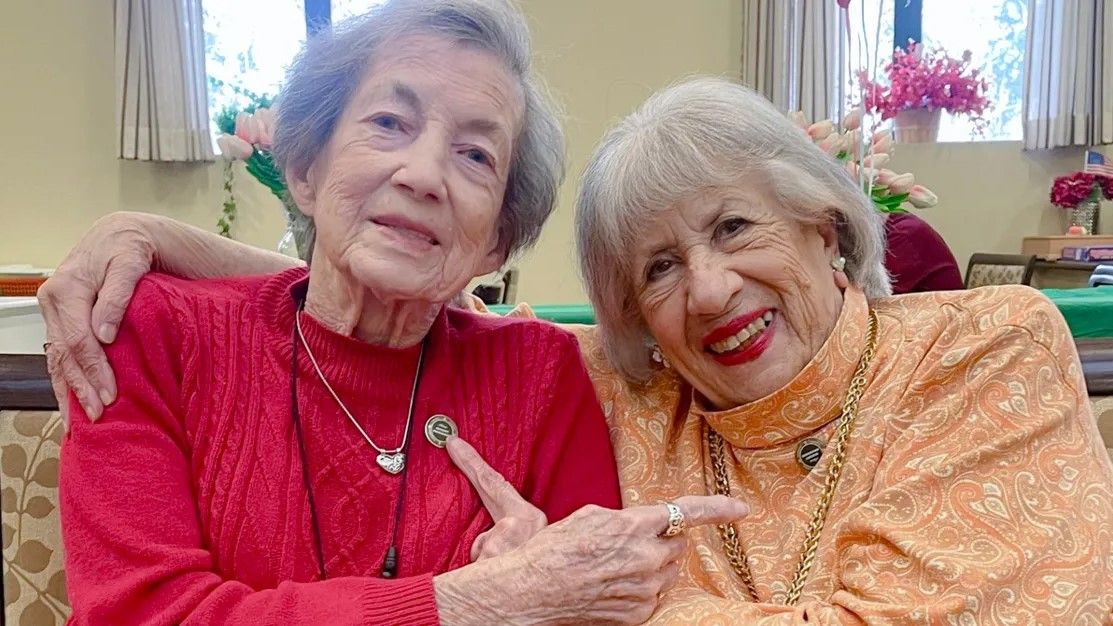 Two older women smiling, sitting closely. One wears a red sweater, the other an orange one. Both point to pins on their chests.