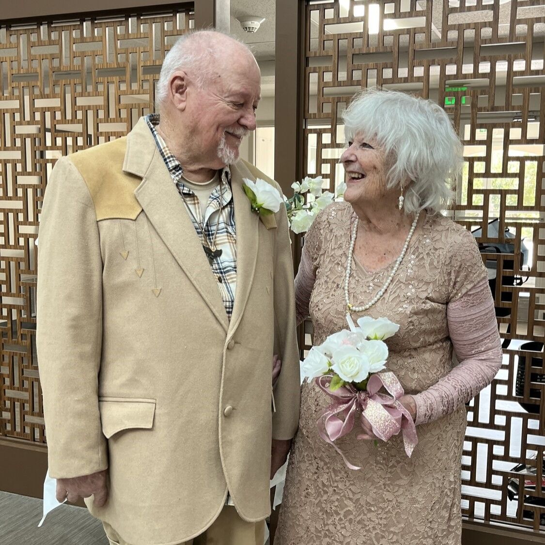 Older couple in formal attire smiling at each other. The woman holds a bouquet of white flowers. They stand in front of a decorative screen.