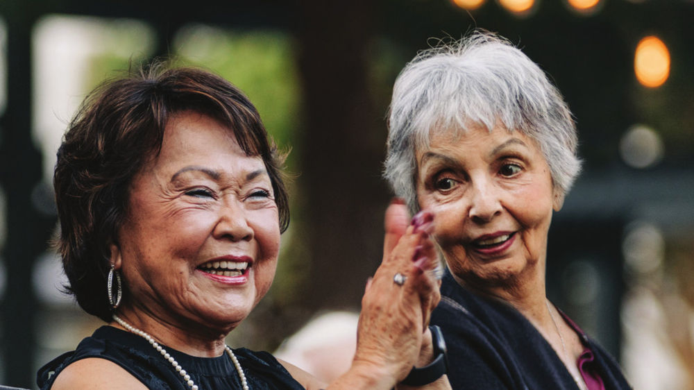 two senior women sitting outdoors on a bench, laughing together