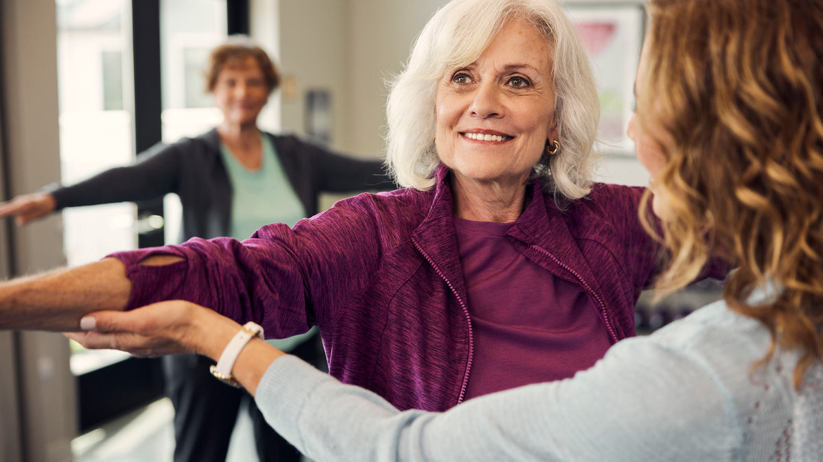 Two women in a fitness class, one assisting the other with arm stretches. Both are smiling and appear engaged in the activity.