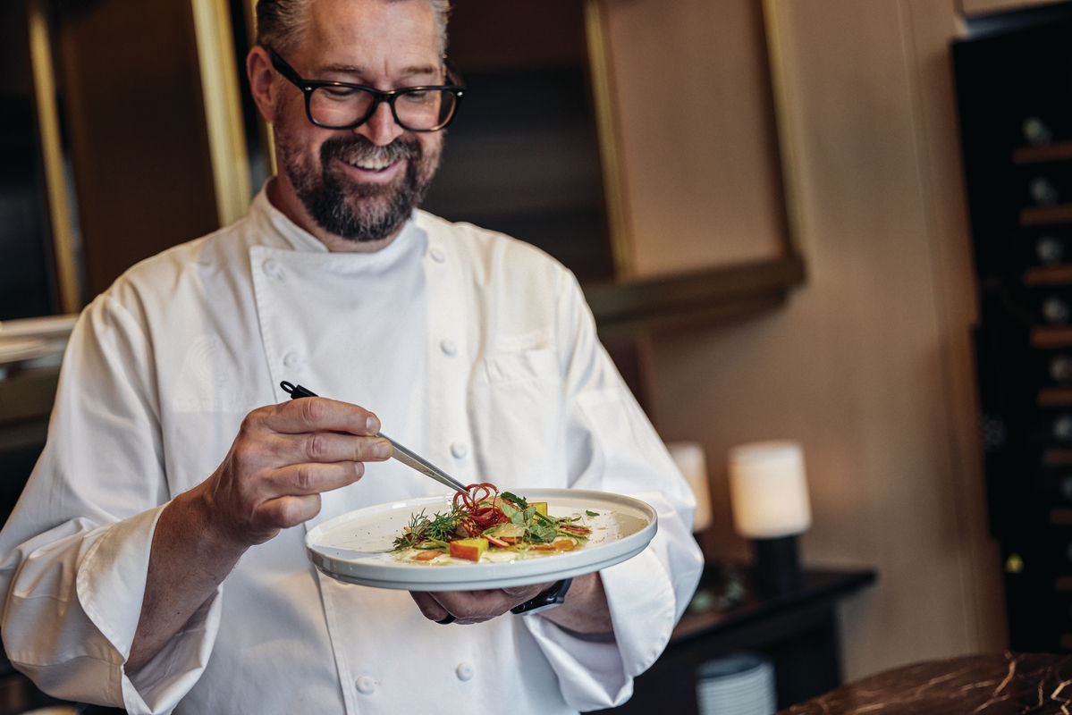 Chef in a white coat smiling while garnishing a dish with herbs and thinly sliced vegetables in a restaurant kitchen.