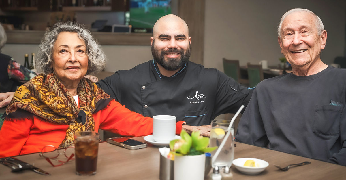 Three people smiling at a dining table, with drinks and utensils in front of them, in a warmly lit restaurant setting.