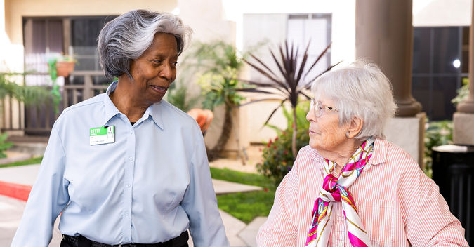 Betty Murrell in a blue shirt assists an older woman with white hair outdoors, both smiling.