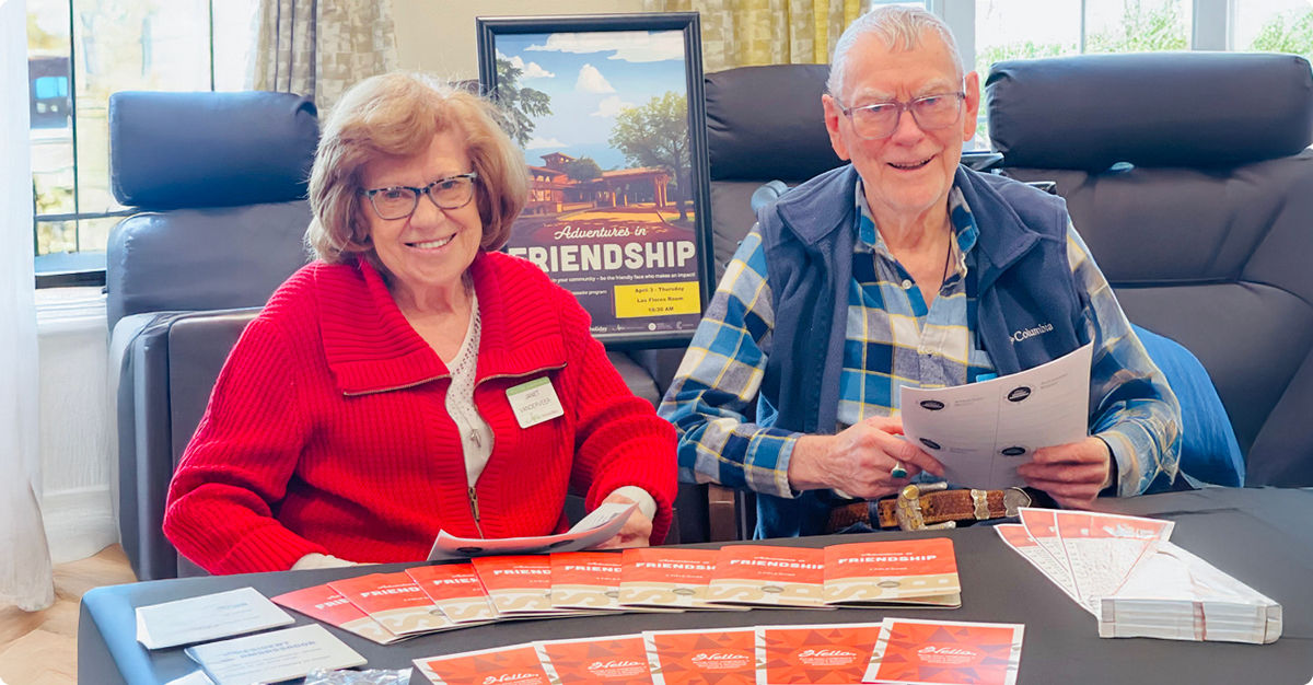 Smiling older couple sitting at a table with brochures titled "Friendship," promoting a community event.
