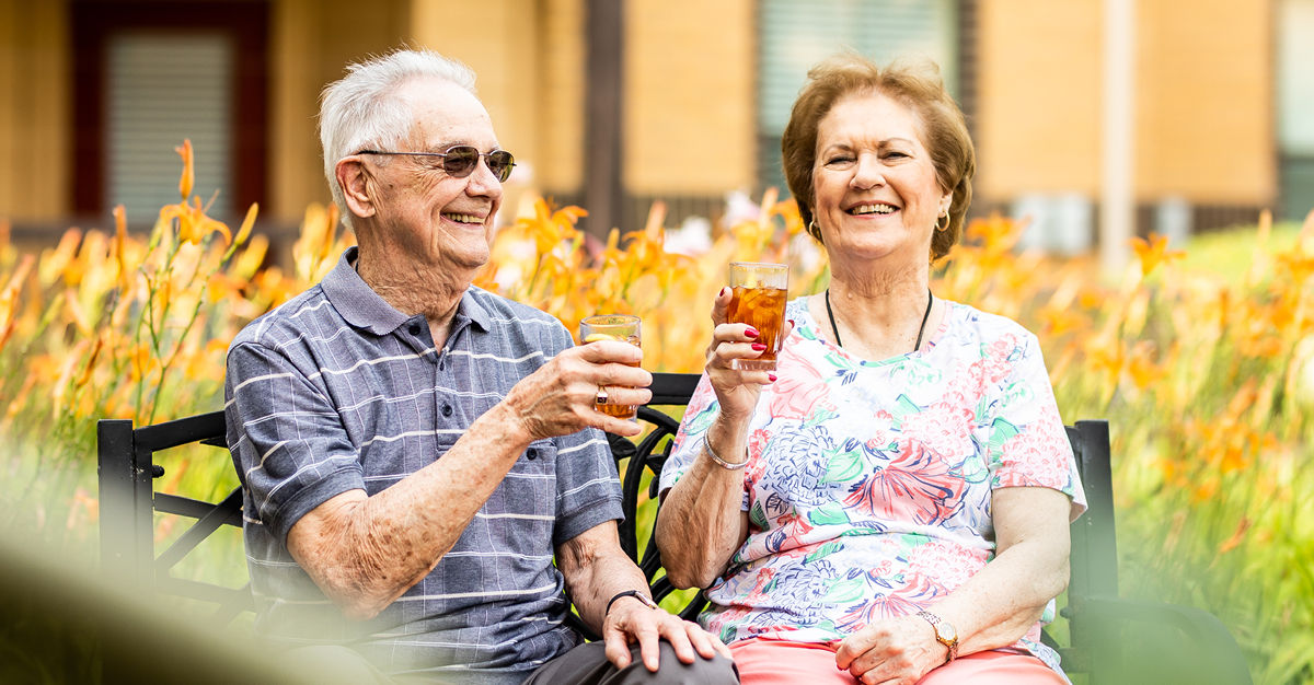 Senior couple sitting on a bench in a garden, smiling and holding glasses of iced tea, surrounded by orange flowers.