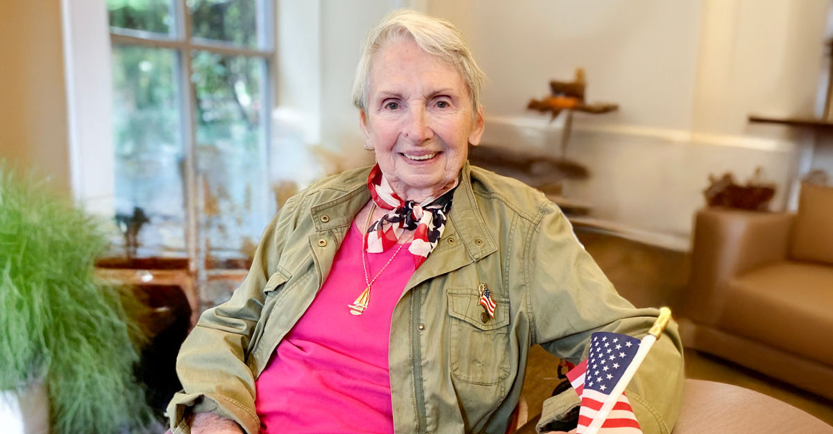 Older adult woman smiling, seated indoors, wearing a green jacket and pink top, holding a small American flag.