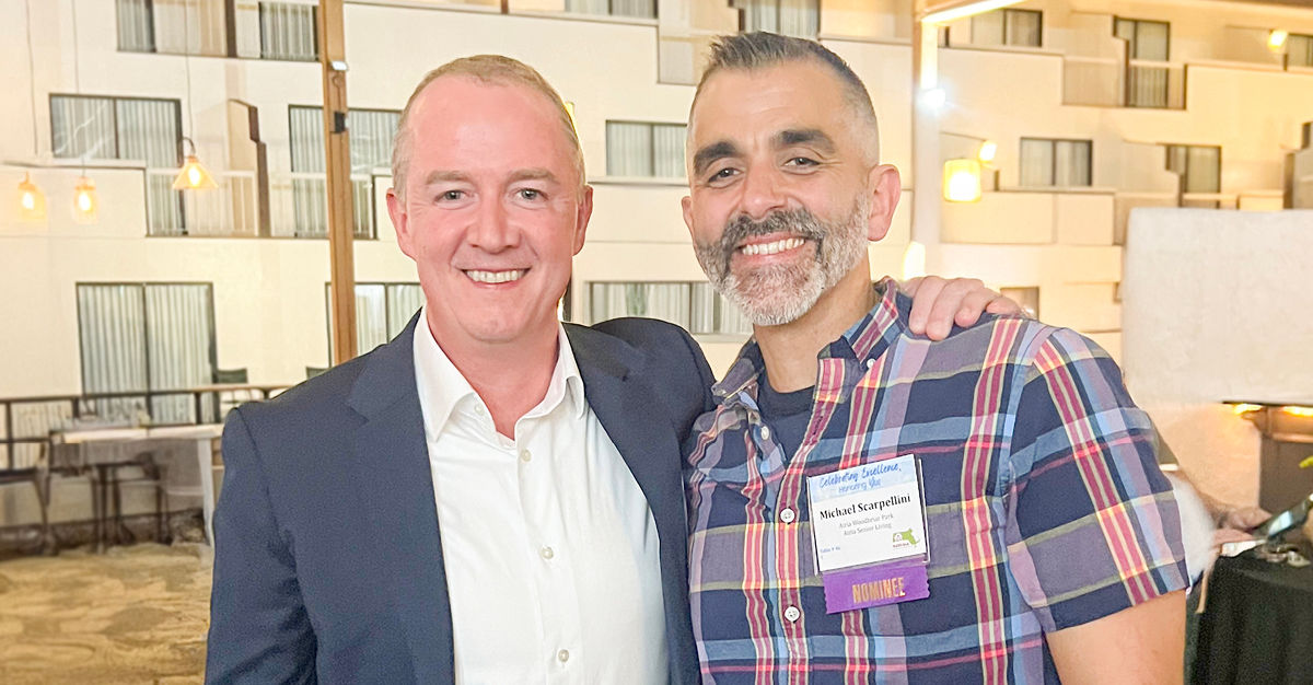 Two men smiling, one in a suit and the other in a plaid shirt, standing indoors with their arms around each other's shoulders.