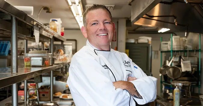 Chef in a white uniform smiling confidently in a well-equipped commercial kitchen.