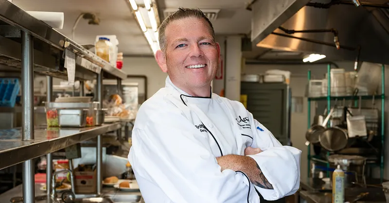 Chef in a white uniform smiling confidently in a well-equipped commercial kitchen.
