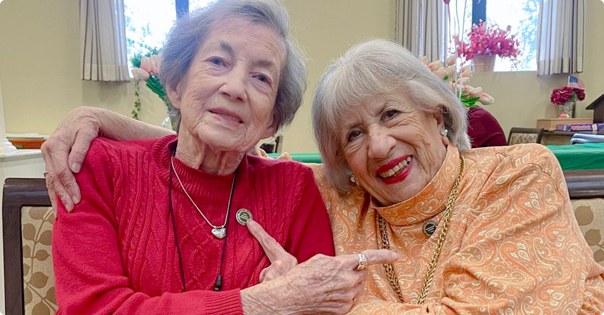 Two older women smiling and pointing at each other, sitting side by side indoors. One wears a red sweater, the other an orange patterned top.