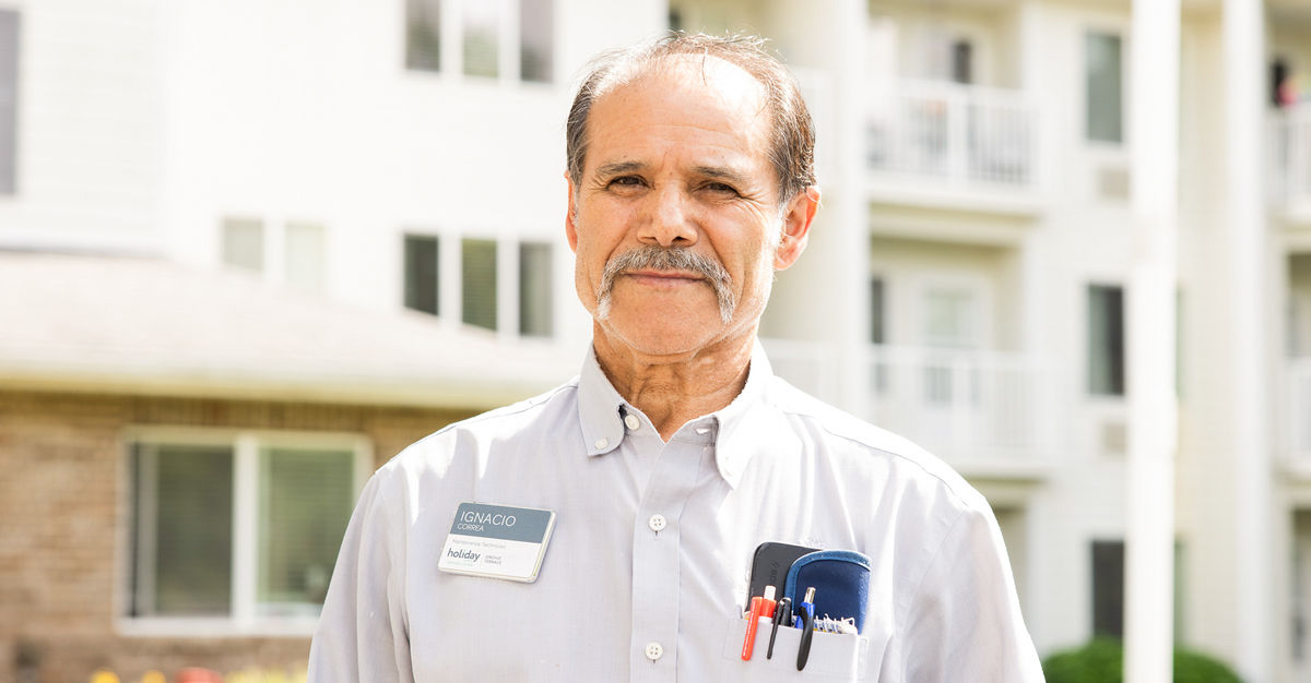 Man with a mustache wearing a name tag and pens in his shirt pocket, standing in front of a building with balconies.