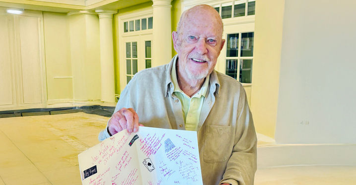 Senior man smiling and holding an open card with handwritten messages, standing in a room with white pillars and large windows.