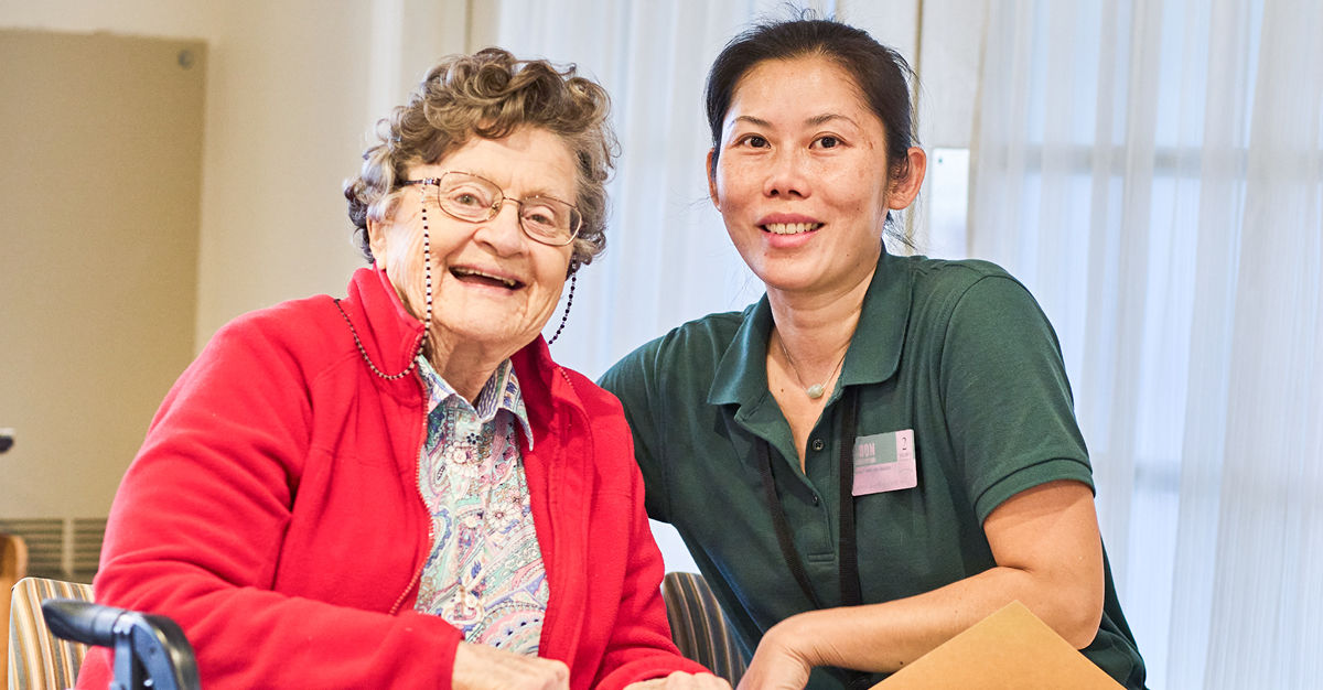 Senior woman in a red jacket smiling, seated next to a caregiver in a green shirt, both looking at the camera in a bright room.