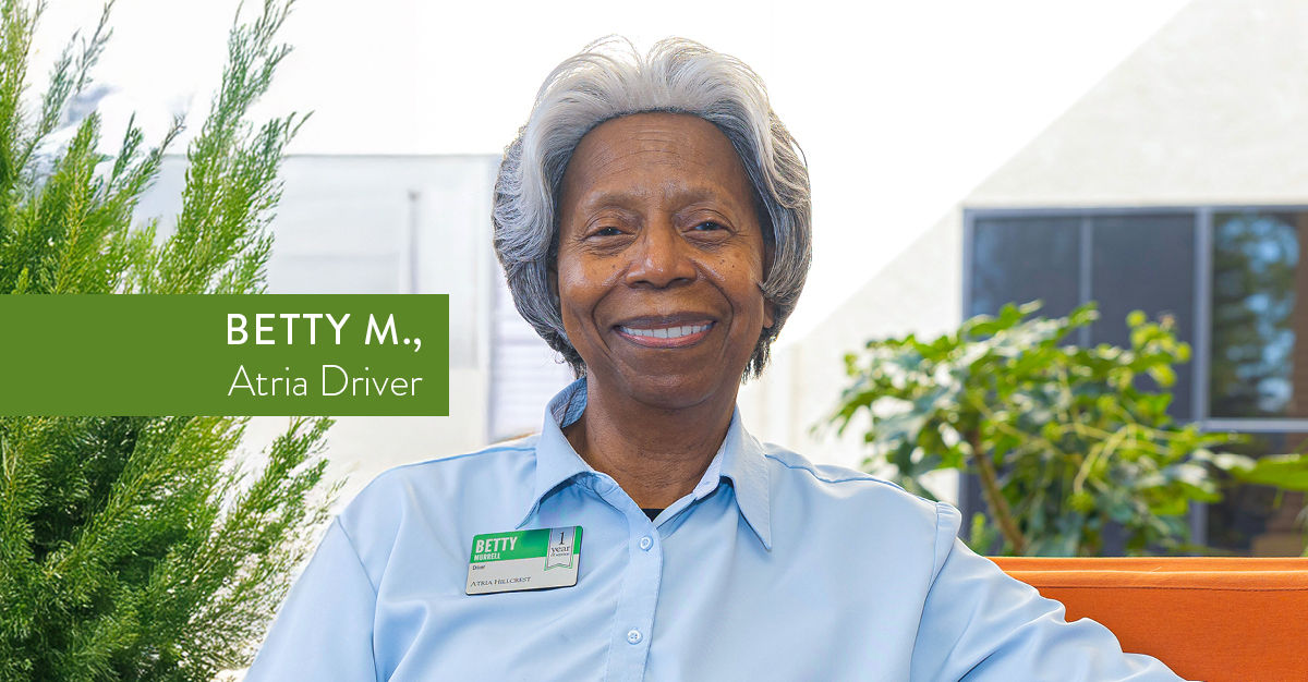 Smiling woman in a blue shirt with a name badge, seated in a bright room with plants. Text reads "Betty M., Atria Driver."