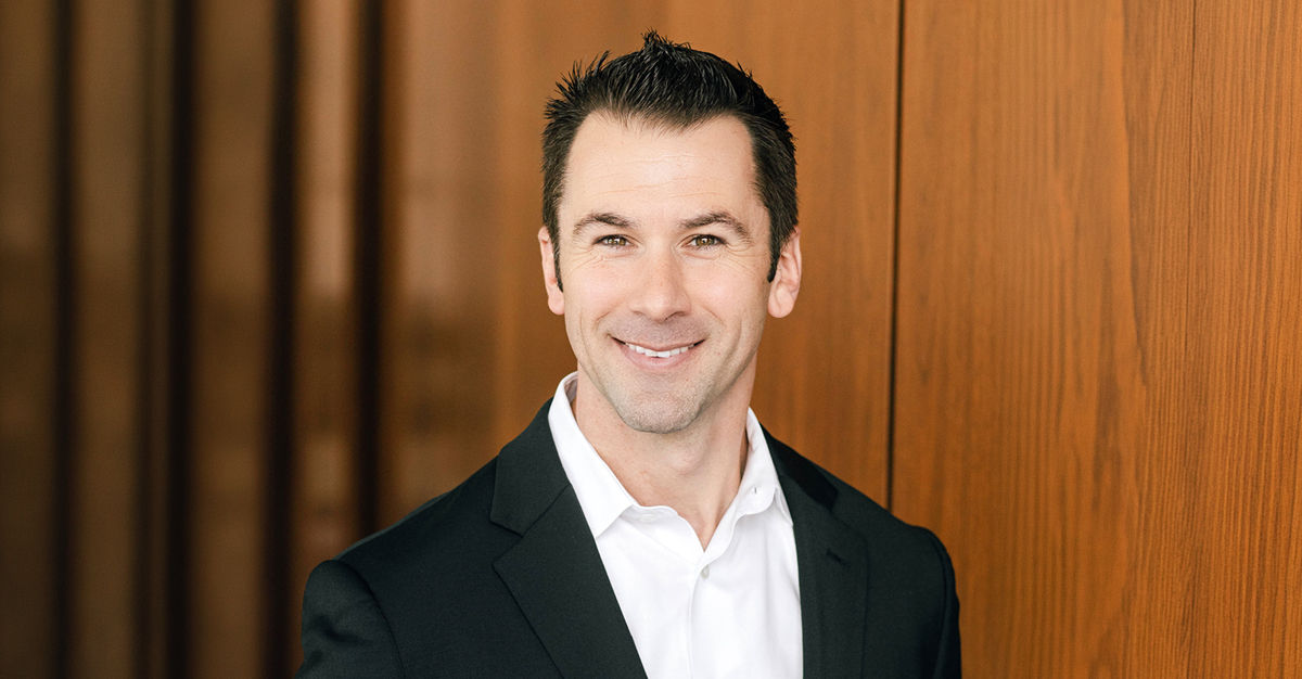 Man in a suit smiling, standing in front of a wooden wall.