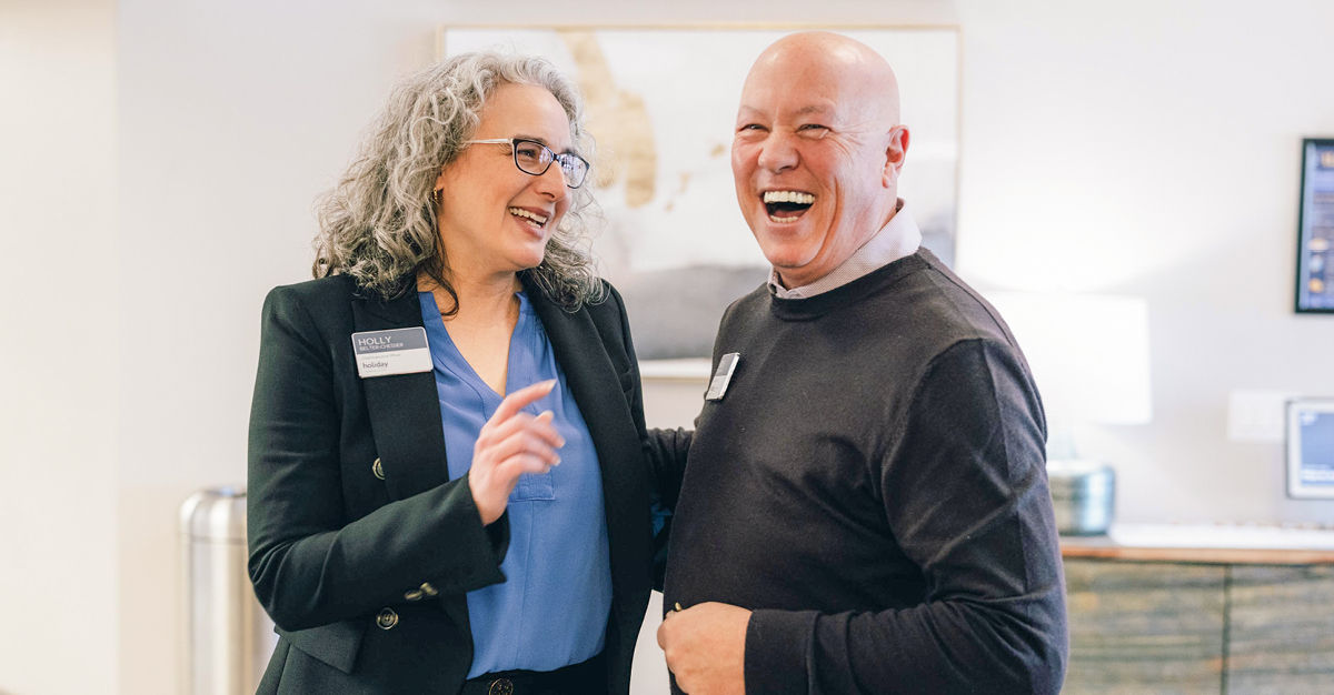 Two people laughing together in an indoor setting, both wearing name tags, with a framed picture and a lamp in the background.