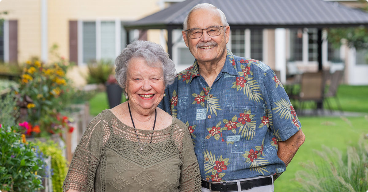 Older couple smiling outdoors, standing in a garden with a gazebo in the background. The man wears a floral shirt; the woman, a green blouse.