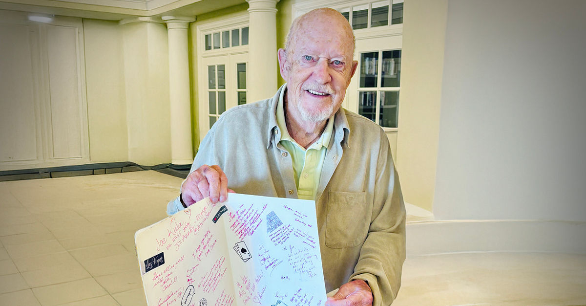Senior man smiling and holding an open card filled with handwritten messages, standing in a bright room with white columns and large windows.