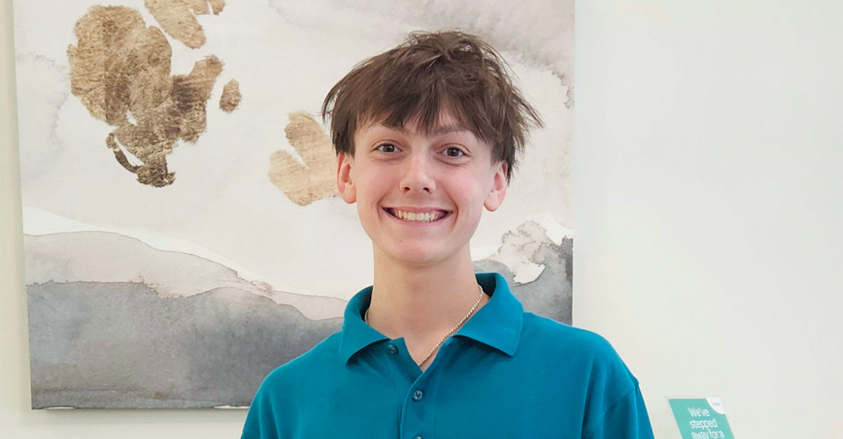 Smiling person with short brown hair wearing a teal shirt, standing in front of a neutral-toned abstract painting.