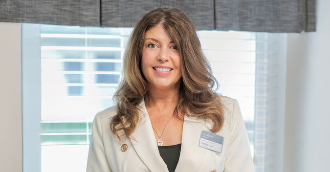 Woman in a white blazer with a name tag, smiling, standing indoors in front of a window with blinds.