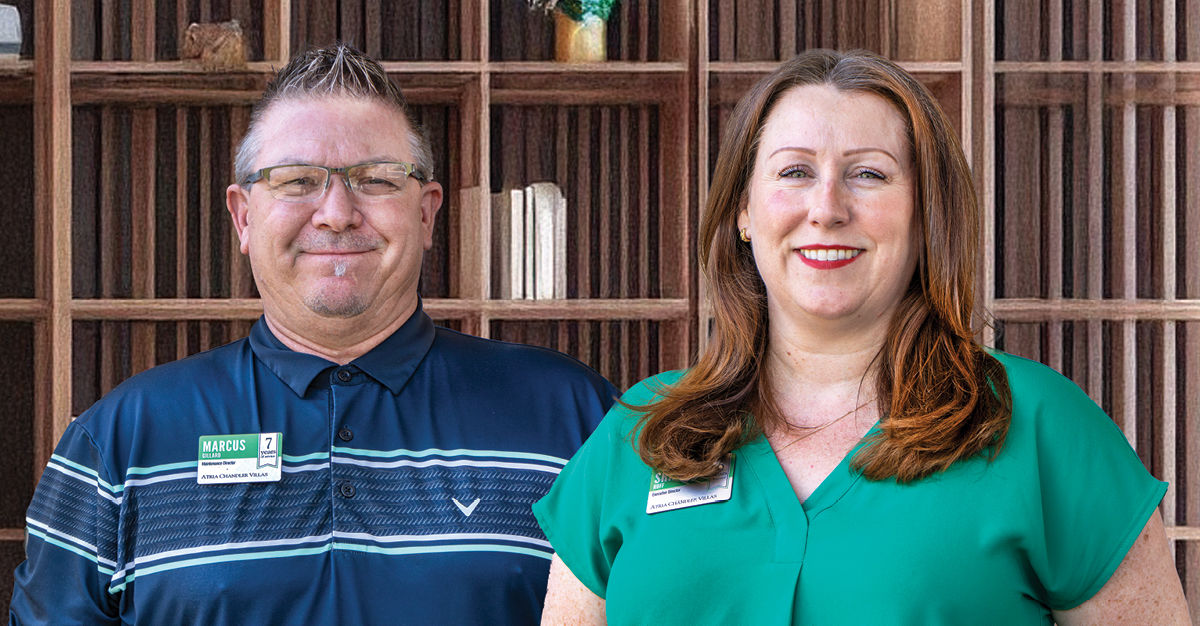 Two people smiling, standing in front of a wooden bookshelf. The man wears glasses and a striped shirt; the woman wears a green blouse.