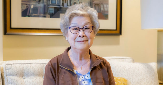 Older woman with glasses and gray hair, smiling while seated on a couch in a warmly lit room with a framed picture behind her.