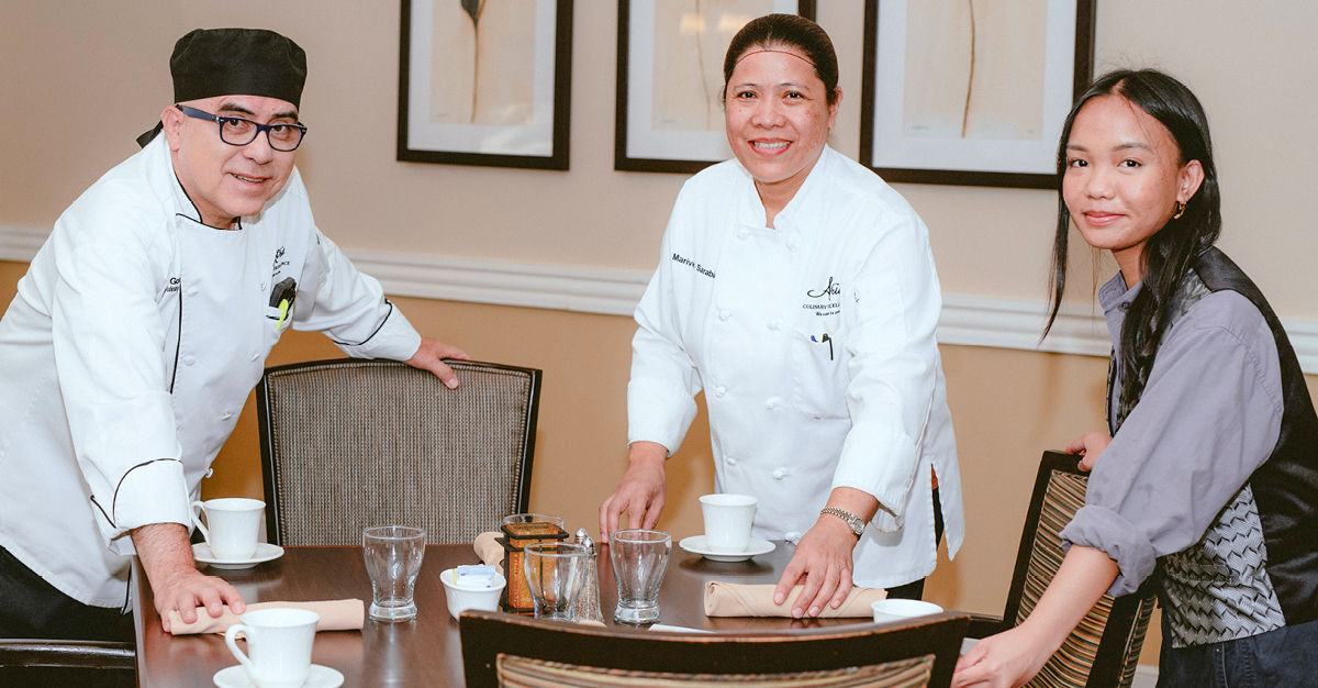Three people, two in chef uniforms and one in casual attire, setting a dining table with cups and glasses in a restaurant.