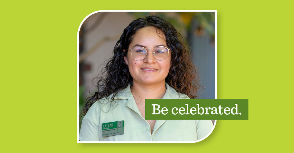 Smiling person with glasses and curly hair in a green shirt, wearing a name tag, with the text "Be celebrated." on a green background.