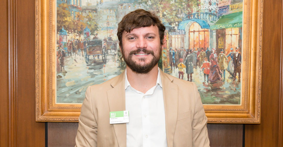 Man with a beard in a tan blazer stands smiling in front of a colorful cityscape painting.